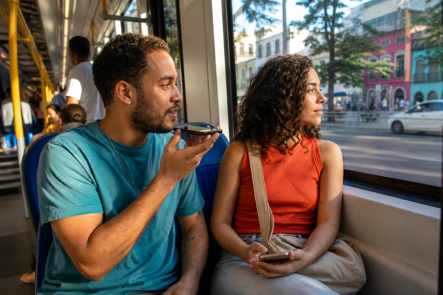 Dois amigos viajam em um ônibus urbano iluminado pelo sol; um grava uma mensagem de áudio e a outra segura o telefone abaixado enquanto observa a cidade pela janela.