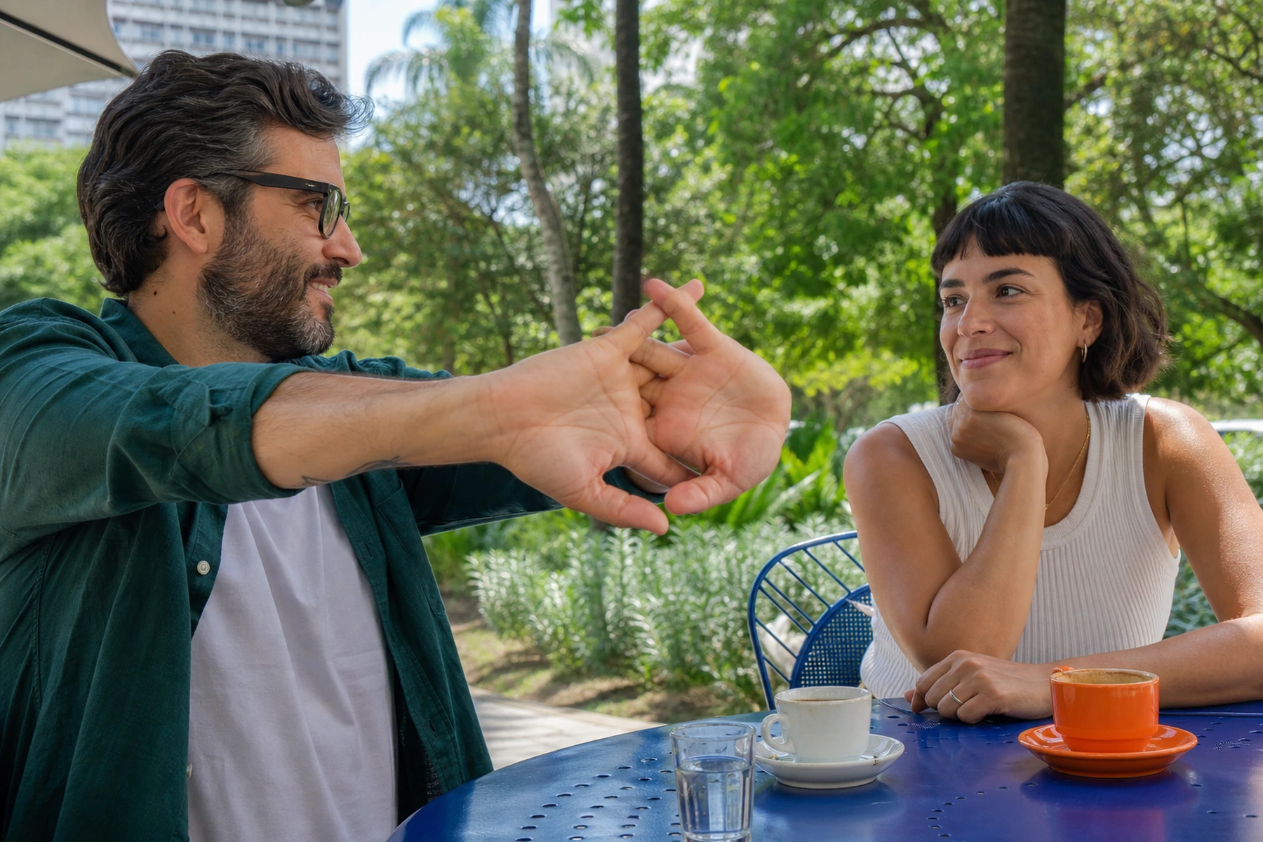 Num café ao lado de um parque, um homem estica as mãos com os dedos entrelaçados enquanto uma amiga observa o gesto com um sorriso discreto.
