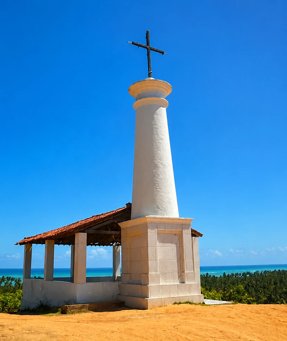 Cruzeiro de alvenaria branca com cruz de ferro no alto de uma barreira de areia, com telhado de telhas ao lado e o mar de recifes ao fundo.