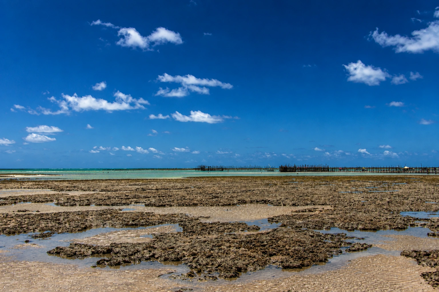 Recife exposto na maré baixa, com poças de água entre as formações e a linha de espuma no horizonte, sob céu azul com nuvens.