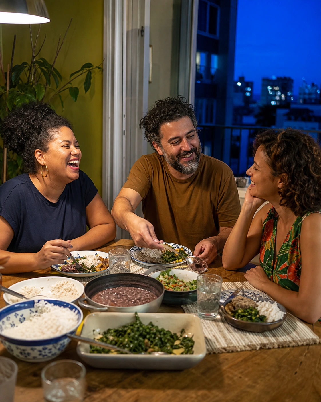 Três amigos conversam e riem em torno de uma mesa com arroz, feijão, verduras e outros pratos, num apartamento iluminado no começo da noite.
